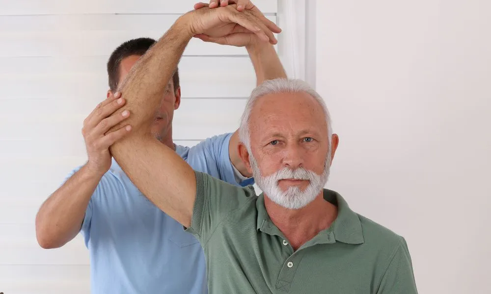 Older man receives physical therapy for lower back pain relief, therapist assists with arm movement.