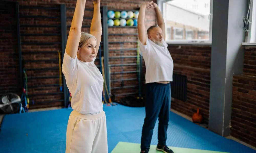 Two older adults stretching in a gym during an exercise physiology session.