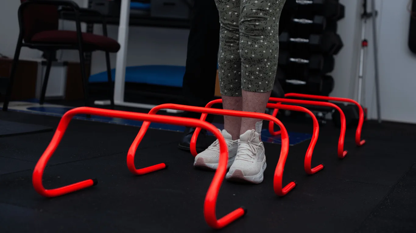Person stepping over small red hurdles in a gym, guided by a physiotherapist.