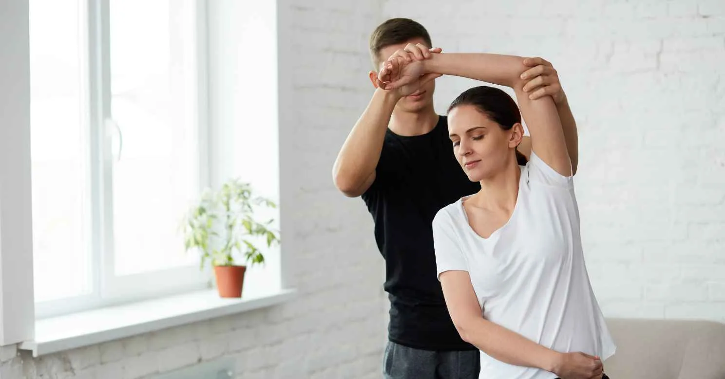 A woman receives shoulder stretching assistance for post-surgery rehab in a bright room.